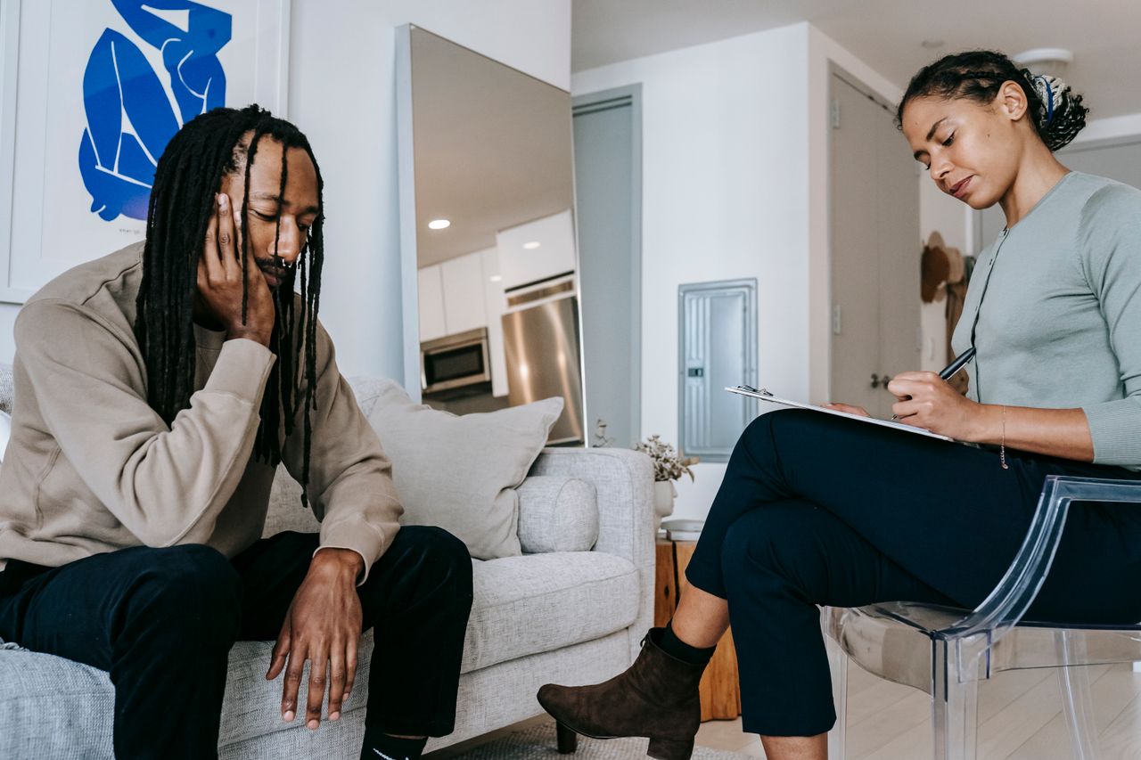 expertise-01 Attentive African American female with clipboard taking notes near sad male partner touching face on couch at home