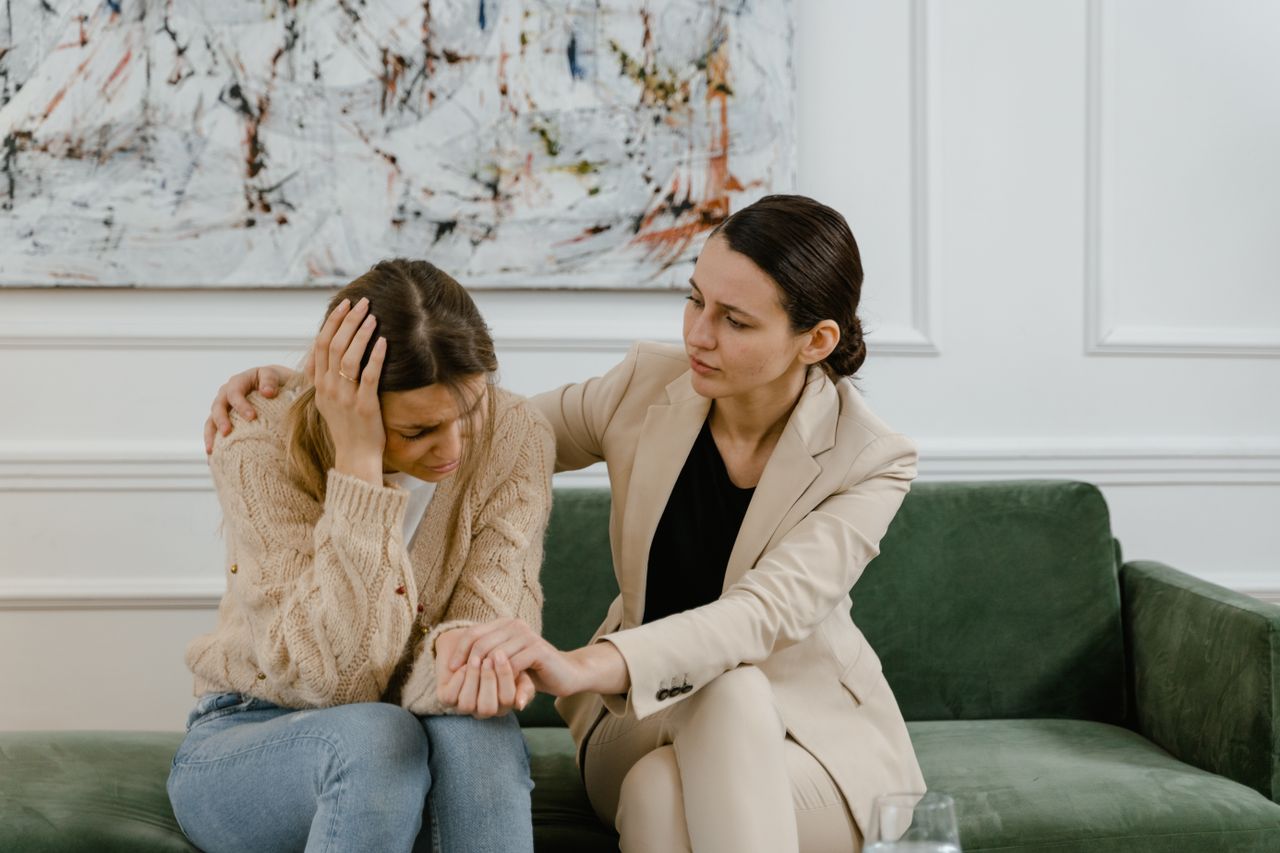 A woman comforts a friend in distress on a green couch indoors.